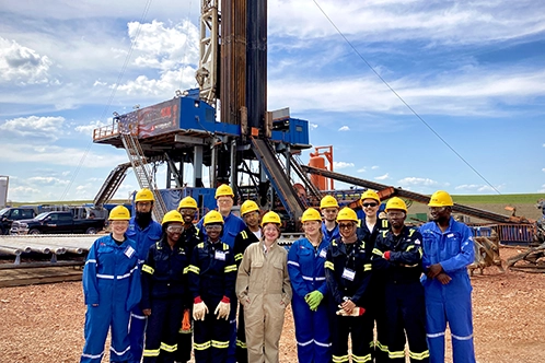 Group of 14 interns pose for a photo in front of an oil rig. They are wearing yellow hard hats and jumpsuits.