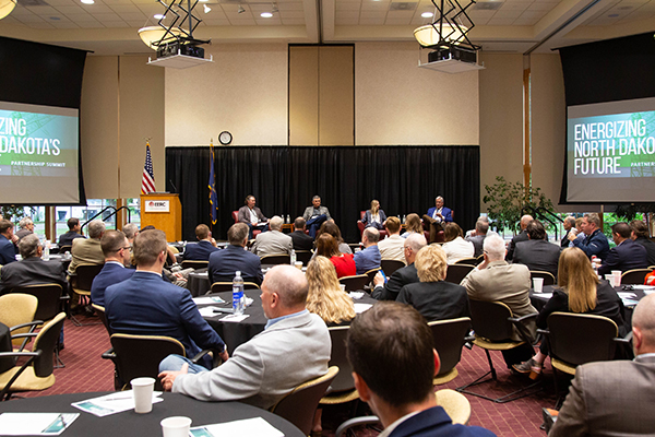 Crowd seated around tables facing a stage with four speakers.